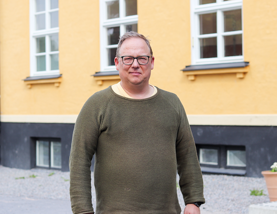 Person står utomhus framför en gul byggnad med vita fönster. Personen bär glasögon och en mörkgrön tröja.