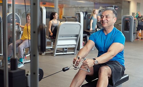 En man tränar på gymmet genom att använda en roddmaskin. Han ler medan han drar i handtaget. I bakgrunden syns andra personer som tränar på olika maskiner.