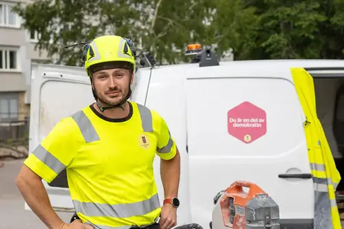 En person i varselkläder och hjälm står framför en vit servicebil. Personen har en neongul tröja med reflexdetaljer och Sundbybergs stads logotyp på bröstet. På sidan av bilen finns en rosa skylt med texten ”Du är en del av demokratin”. I förgrunden syns arbetsutrustning, och i bakgrunden finns träd och bostadshus.