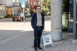 En man står på en trottoar i en stadsmiljö. Han bär glasögon, mörkblå kofta, grå skjorta, mörka byxor och svarta skor. I bakgrunden syns flerbostadshus, parkerade bilar och träd. Bredvid mannen står en vit skylt med texten ”Här kan du Förtidsrösta” och en pil som pekar åt höger.